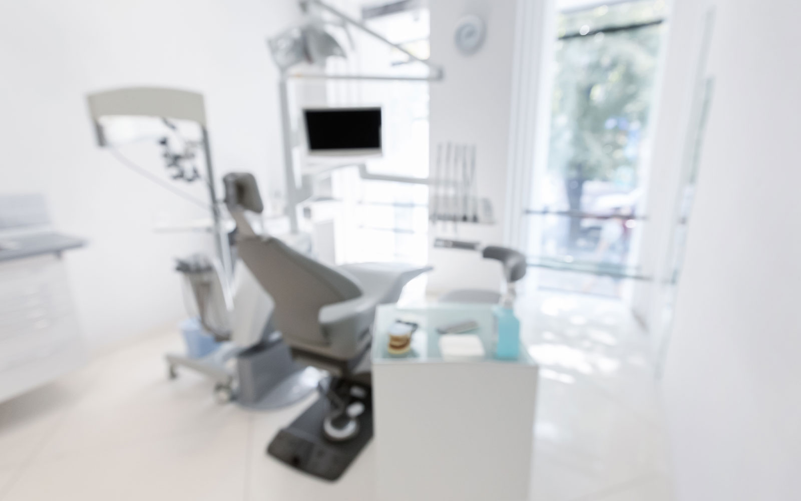 The image depicts a modern dental office interior with a large chair, a dental examination table, and various equipment.