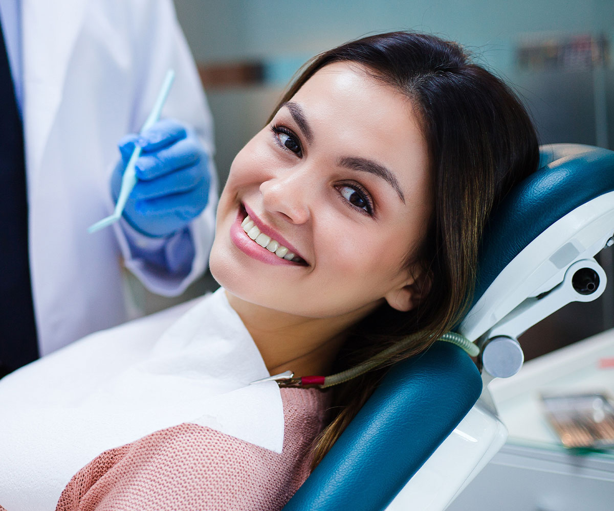 The image shows a smiling woman sitting in a dental chair, receiving dental care from a professional wearing a blue surgical gown and a white face mask.